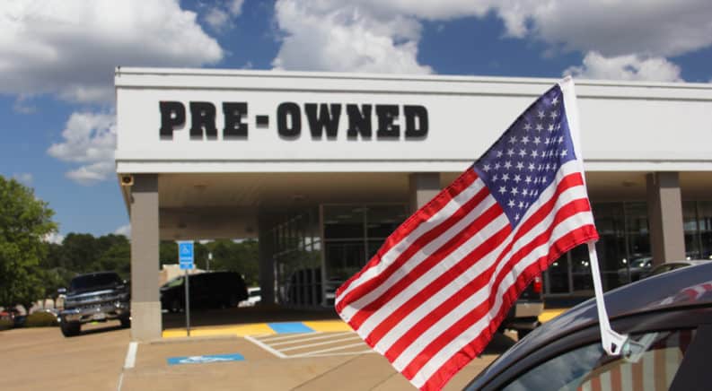 American flag on a vehicle in front of a white building with the word "Pre-Owned" on it
