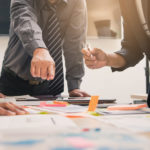 Three men and one woman in business clothing standing around a brown table covered in papers and sticky notes