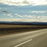 Empty grey road with large brown field, mountains, and a blue sky with white clouds in the background