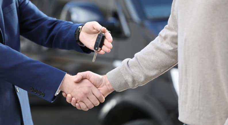 Man in a blue suit shaking hands with and handing a car key to a man in a tan sweater