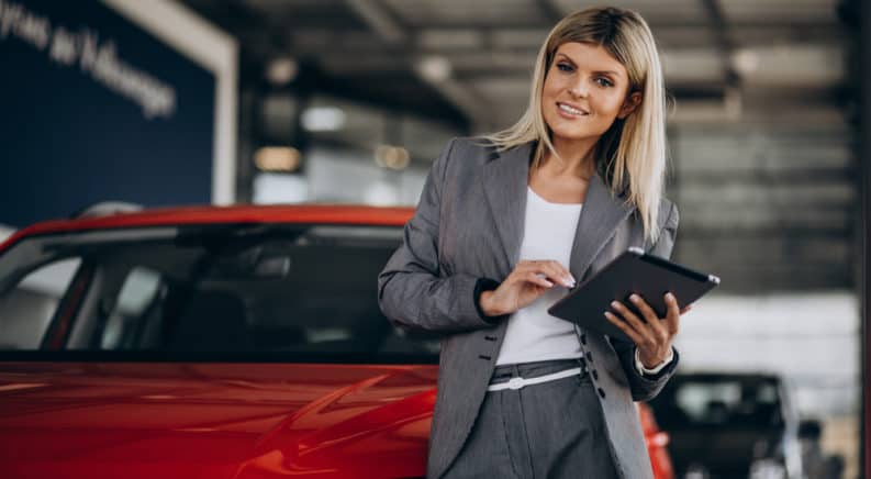 Woman in a grey suit and white shirt leaning against a red car and using a tablet