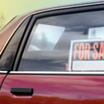 Red car with a black, orange, and white "For Sale" sign in the window