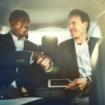 Two diverse businessman smiling and shaking hands together while sitting in the backseat of a car driving through the city talking about cadillac lease deals.