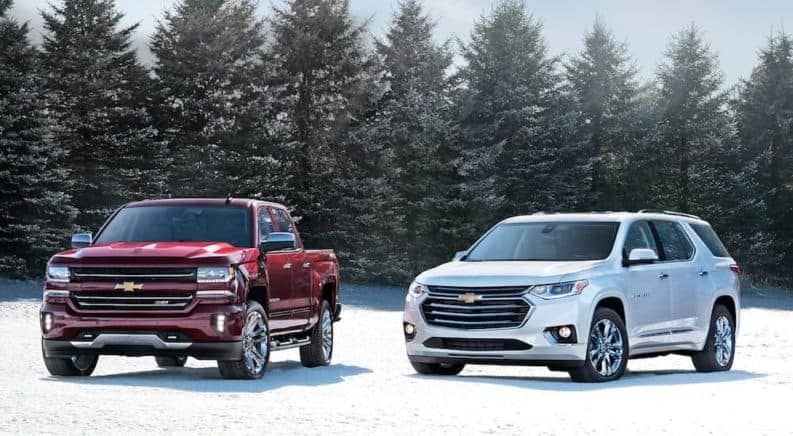 Red 2019 Chevy Silverado and white 2019 Chevy Traverse in snowy field with evergreen trees in back