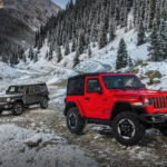 A red two door 2018 Jeep Wrangler is in front of a silver four door Jeep Wrangler hard top on a snowy trail with mountains behind them.