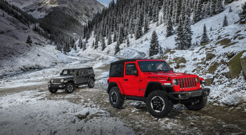 A red two door 2018 Jeep Wrangler is in front of a silver four door Jeep Wrangler hard top on a snowy trail with mountains behind them.