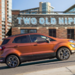 An orange 2019 Ford Ecosport is driving up a city block on a sunny day.
