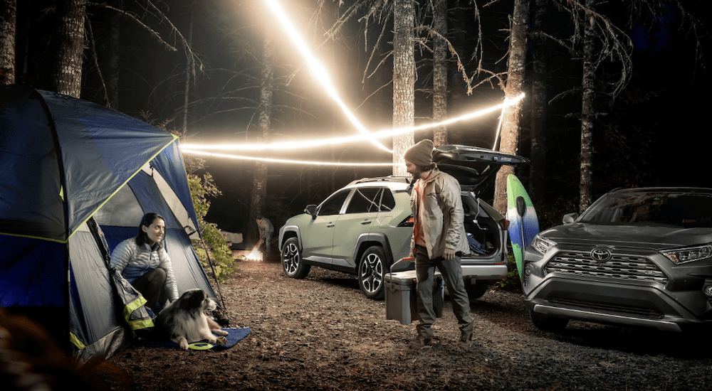 A man is unloading camping gear from his green 2019 Toyota Rav 4 which is parked next to a grey one. The campsite is dark with string lights set up. Find the Rav 4 at Toyota Dealership