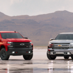 A red 2019 Chevy Silverado with black trim is next to a silver 2019 Silverado in front of desert mountains.
