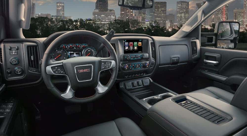 The black interior of a 2019 GMC Sierra 2500 HD is shown with a city skyline through the windshield. Compare interior features when looking at the 2019 GMC Sierra 2500HD vs 2019 Ford F-250.