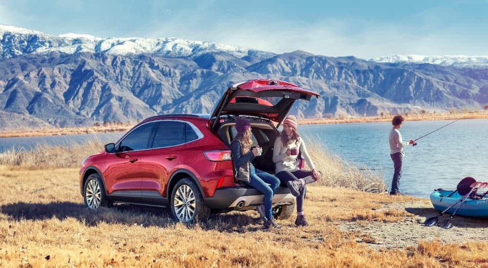 Ford's future 2020 Escape will feature hybrid options. This red Escape shows two people sitting on the rear bumper in front of mountains while another person fishes.