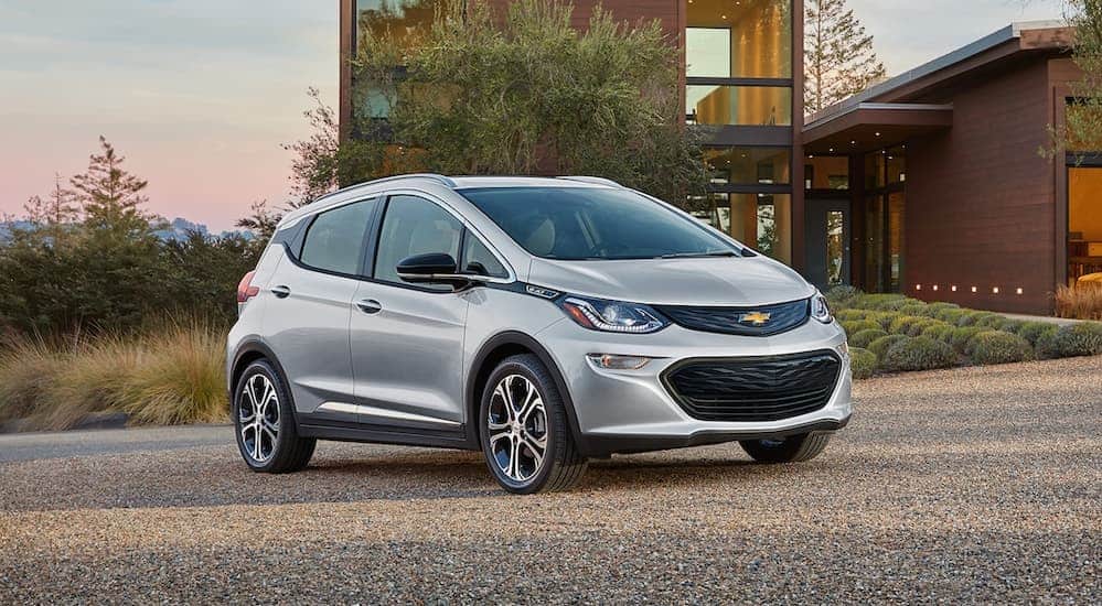 A silver 2019 Chevy Bolt is parked in the driveway of a modern house. It is one of the current Chevy cars offered.