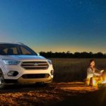 A white 2019 Ford Escape is parked next to a couple at a campfire at night.