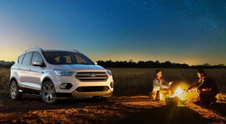 A white 2019 Ford Escape is parked next to a couple at a campfire at night.