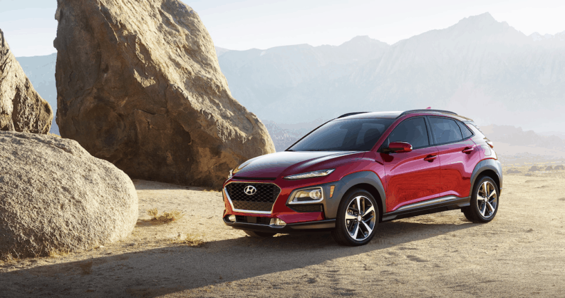 A red 2019 Hyundai Kona is parked next to a rock formation in the desert.