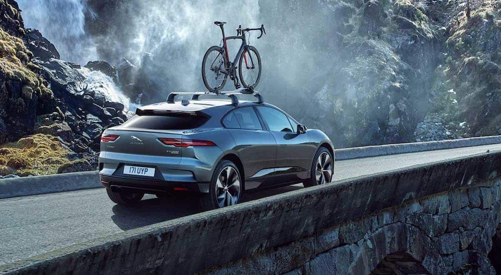 A grey 2019 Jaguar I-Pace is driving over a stone bridge next to a waterfall with a bike on the roof.