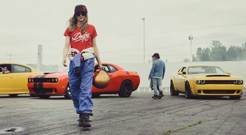 A girl with a vintage look is walking away from two 2019 Dodge Challengers and a yellow Charger on a track.