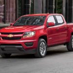 A red 2019 Chevy Colorado drives through a downtown. The Colorado is the smallest of the Chevy trucks for sale.