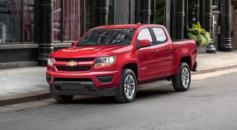 A red 2019 Chevy Colorado drives through a downtown. The Colorado is the smallest of the Chevy trucks for sale.