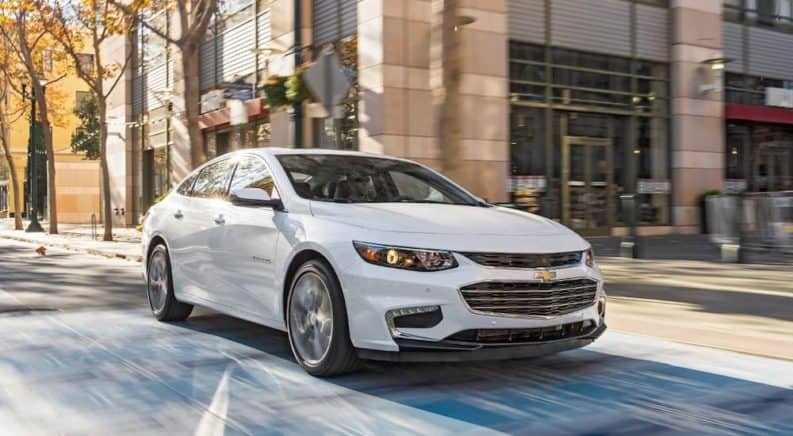A white 2018 Chevy Malibu is driving down a sunny town street.