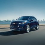 A blue 2018 Chevy Trax, one of the popular used Chevy SUVs for sale, is driving past water with a city skyline in the distance.