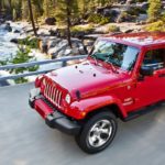 A red 2016 Jeep Wrangler Unlimited is driving on a bridge over a river in the woods.