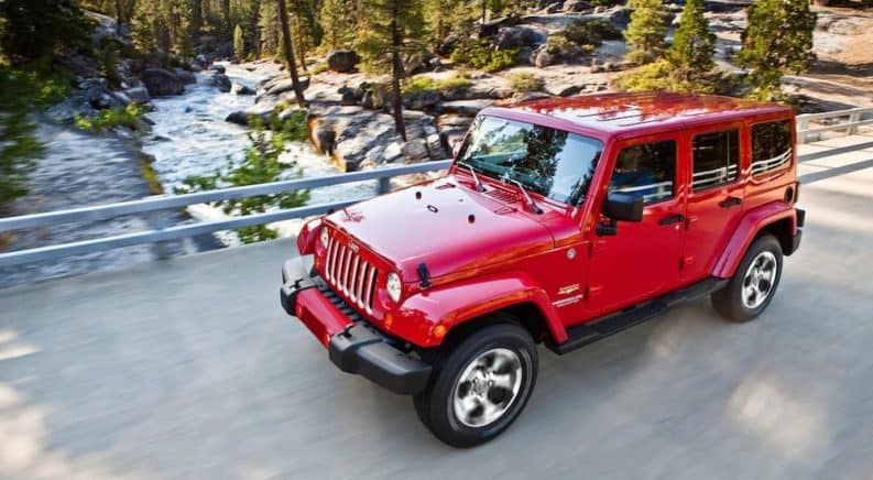 A red 2016 Jeep Wrangler Unlimited is driving on a bridge over a river in the woods.