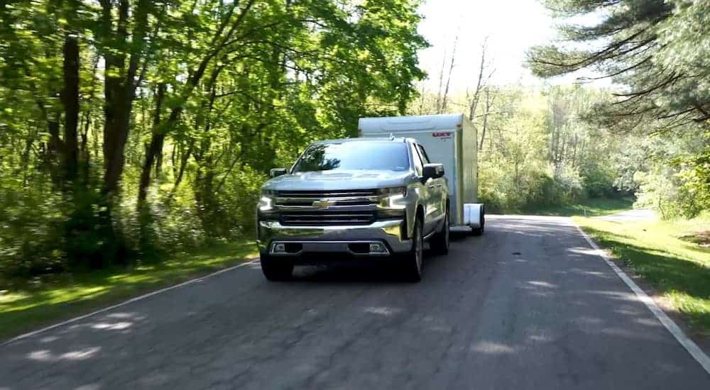 A white 2019 Chevy Silverado 1500 is towing a white trailer on a tree-lined road.