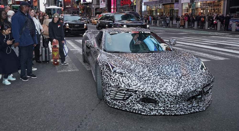 A camouflaged 2020 Chevy Corvette is taking a corner in Time Square with pedestrians looking on.