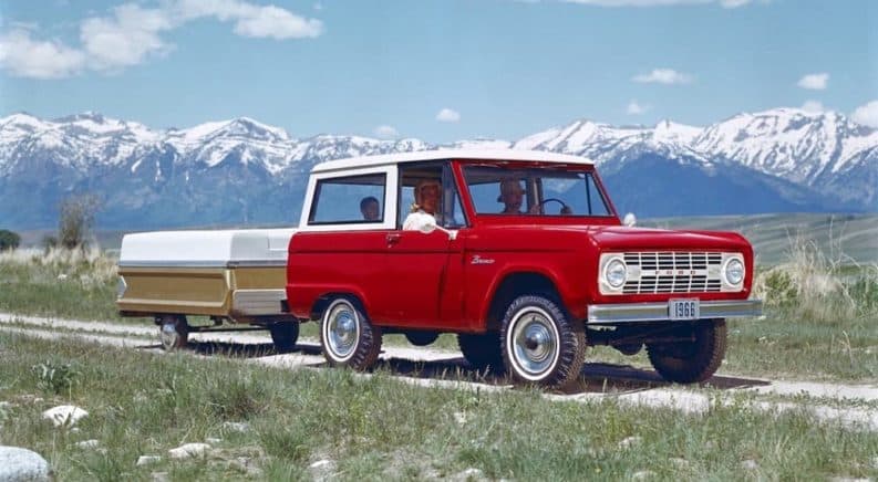 A red 1966 Ford Bronco is towing a popup camper in front of mountains.