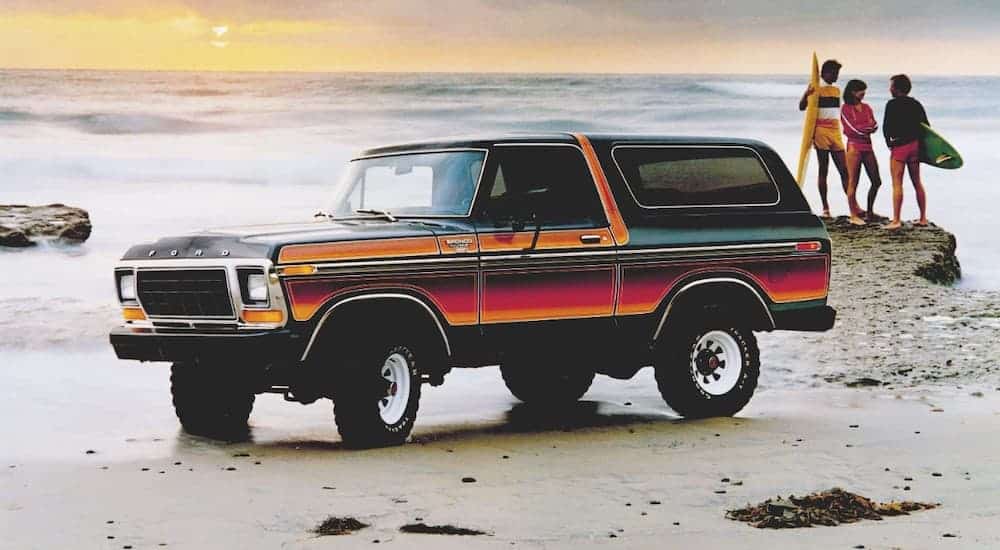 A multi colored 1979 Ford Bronco is on the beach next to surfers.