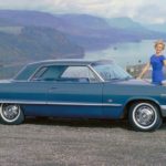 A 1960s woman in a blue dress is next to a blue 1963 Chevrolet Impala overlooking mountains and a river.