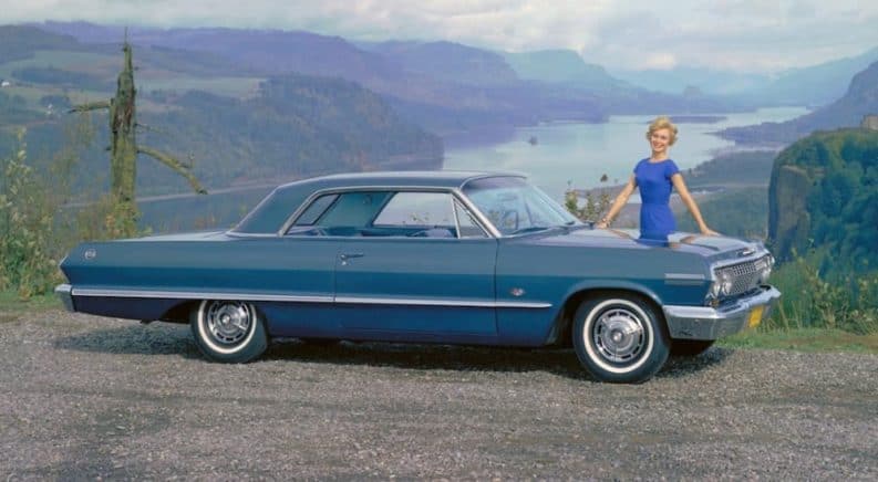 A 1960s woman in a blue dress is next to a blue 1963 Chevrolet Impala overlooking mountains and a river.