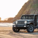 A grey 2018 Jeep Wrangler, a popular Jeep for sale, is on the beach next to a surfer.