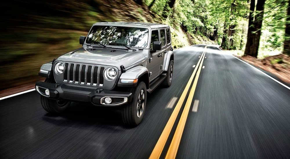 A silver 2019 Jeep Wrangler is driving on a road through the woods.