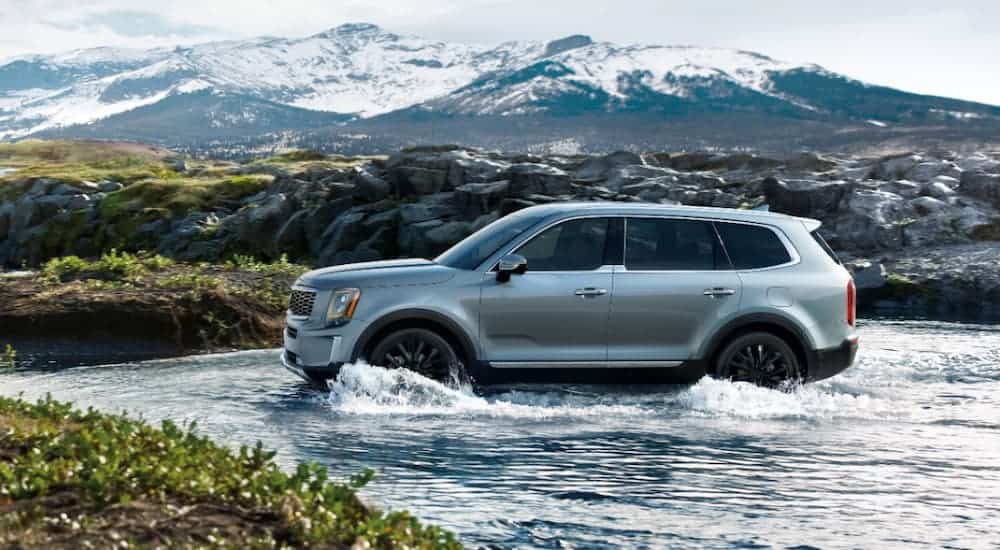 A silvery blue 2020 Kia Telluride is crossing water with white cap mountains behind it.