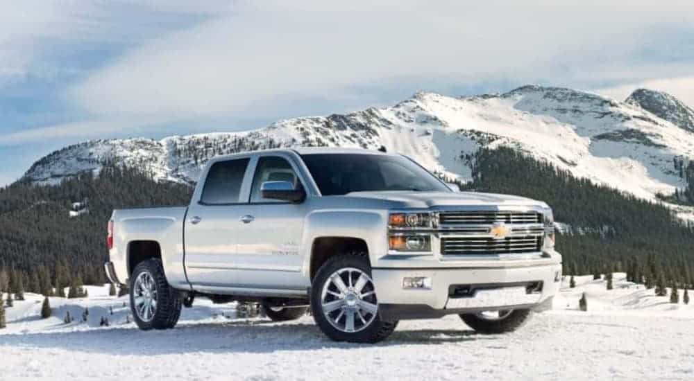 A white 2014 Chevy Silverado, a popular choice for used trucks for sale, is parked in the snow in front of mountains.