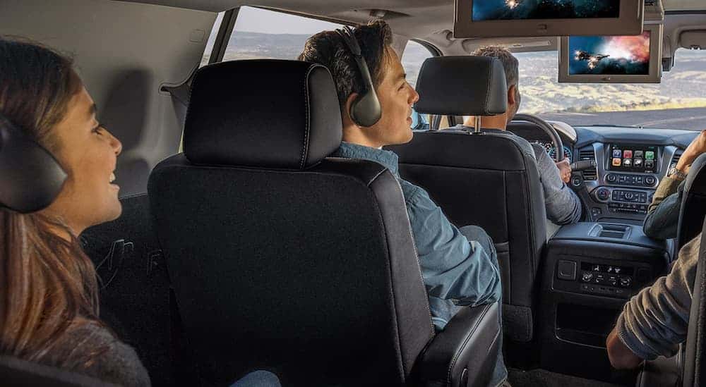 A smiling family is watching a movie on the 2 screens inside the 2020 Suburban. 