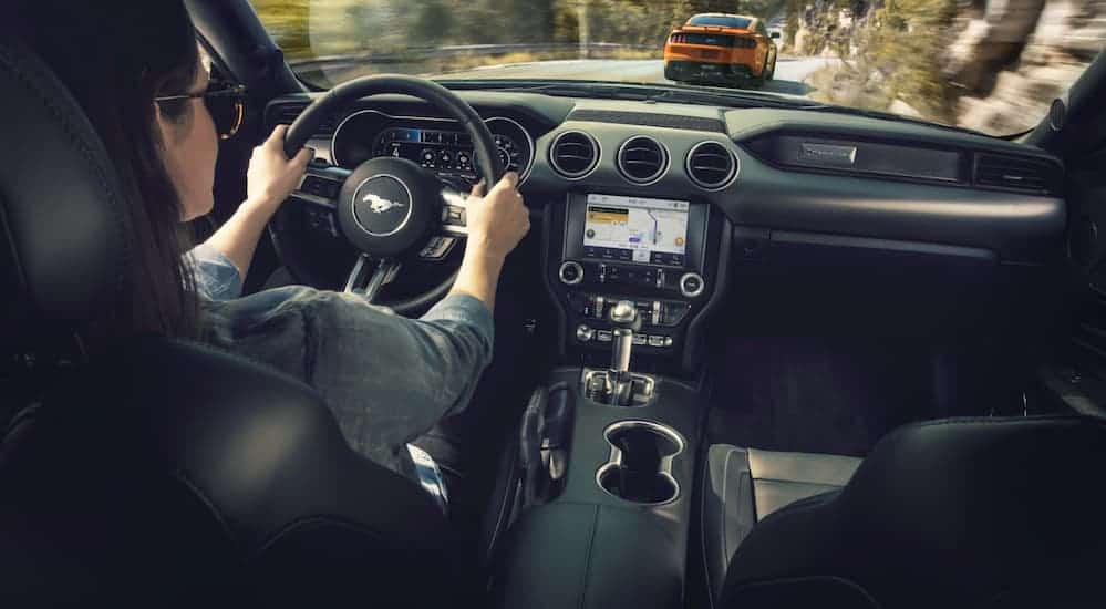 An interior view is shown of a woman driving a 2020 Ford Mustang behind an orange Mustang on a mountain road.