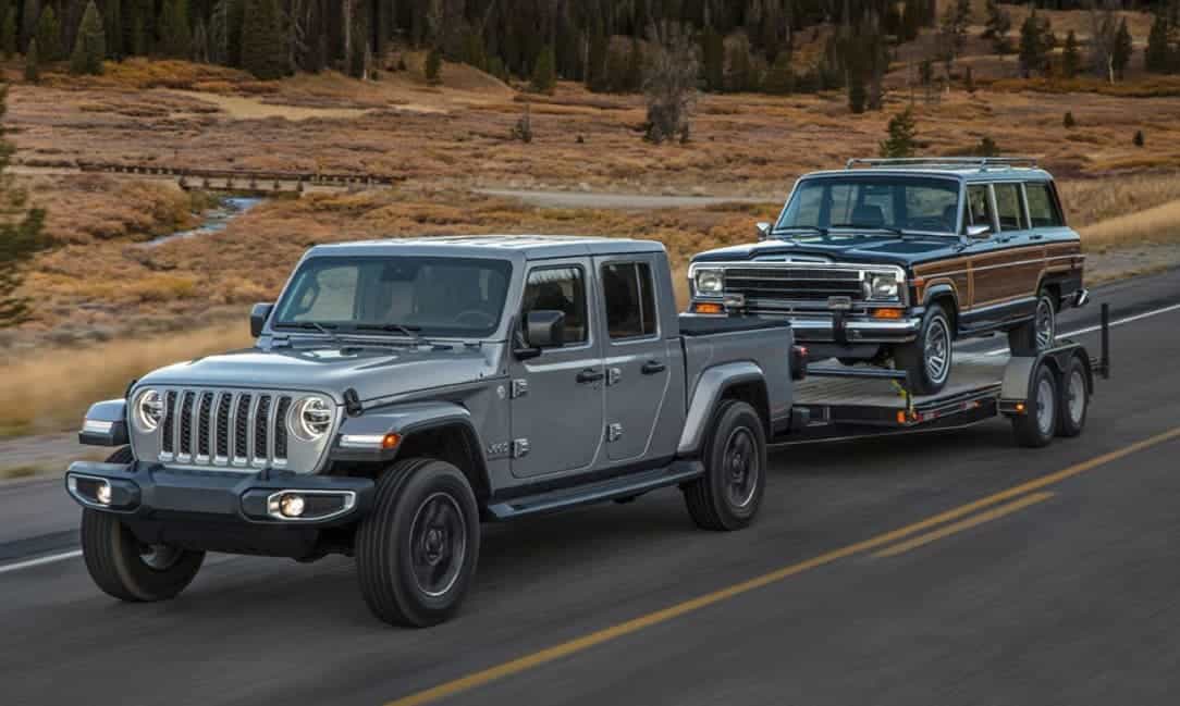 A silver 2020 Jeep Gladiator is towing a wood paneled 1980s Wagoneer down a highway. 