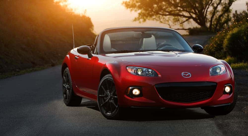 A red 2015 Mazda MX-5 Miata is parked on a treelined road with the sun setting behind it. 