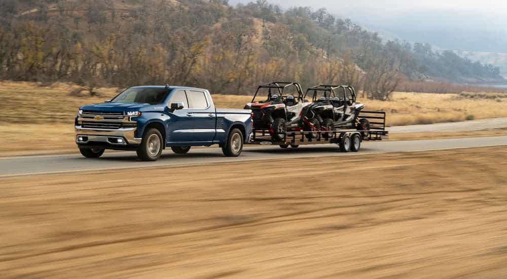 A blue 2020 Chevy Silverado 1500 Diesel is towing 2 side by sides on a large trailer on a grass lined road. 