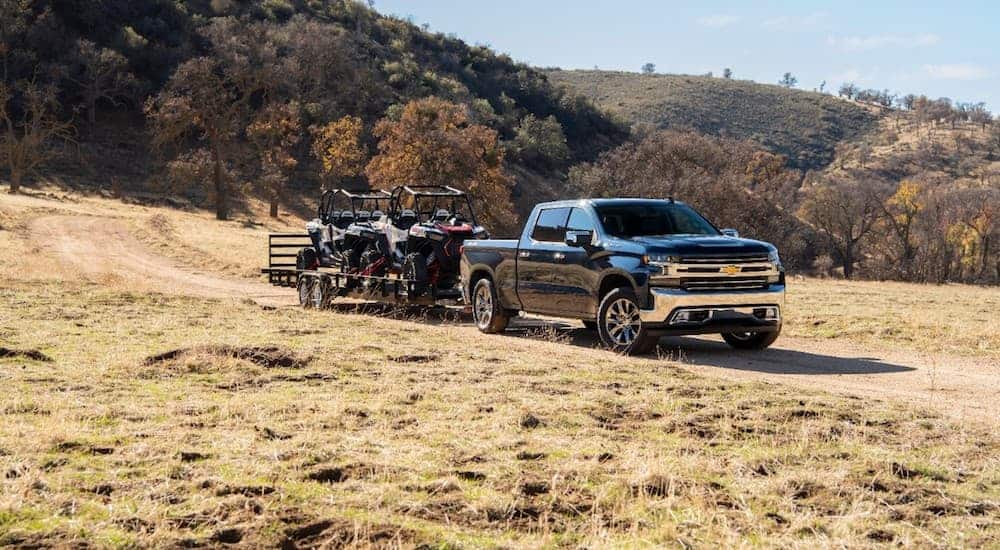A blue 2020 Chevy Silverado 1500 is towing 2 side by sides on a dirt road.