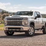 A white 2020 Chevy Silverado HD is parked in a dirt parking lot with grassy mountains in the background.