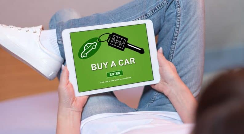 A woman is sitting on her couch while shopping for a new car online on a tablet.