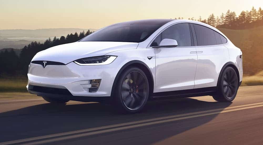 A white Tesla Model X is driving on a road with the sun setting behind it. 