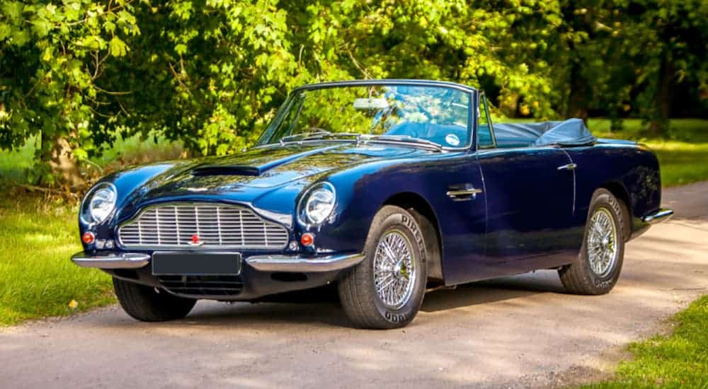 A blue 1968 Aston Martin DB6 Volante is parked next to a tree in the sun. 