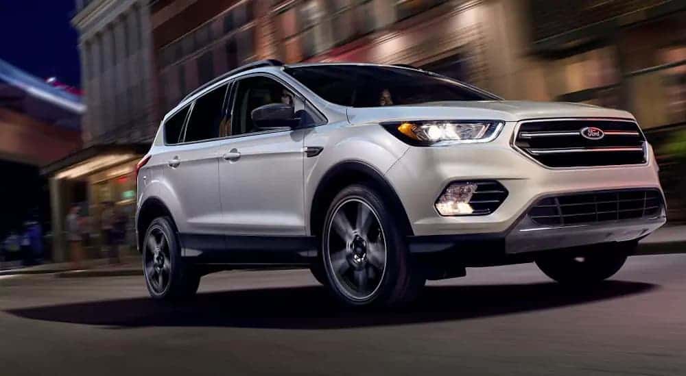A white 2019 Ford Escape is driving on a dark lit city street past blurred buildings. 