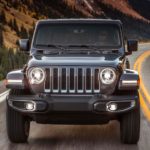 A grey 2020 Jeep Wrangler is driving on a road with snow covered mountains in the distance.