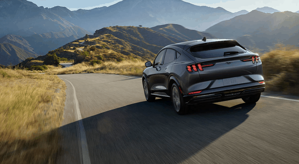 A grey 2021 Ford Mustang Mach-E is driving on a grass lined road towards mountains that are in the distance. 
